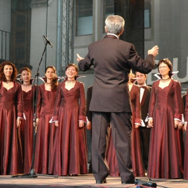 Dirigent vor einem Frauenchor. Die Frauen tragen festliche dunkelrote Kleider.