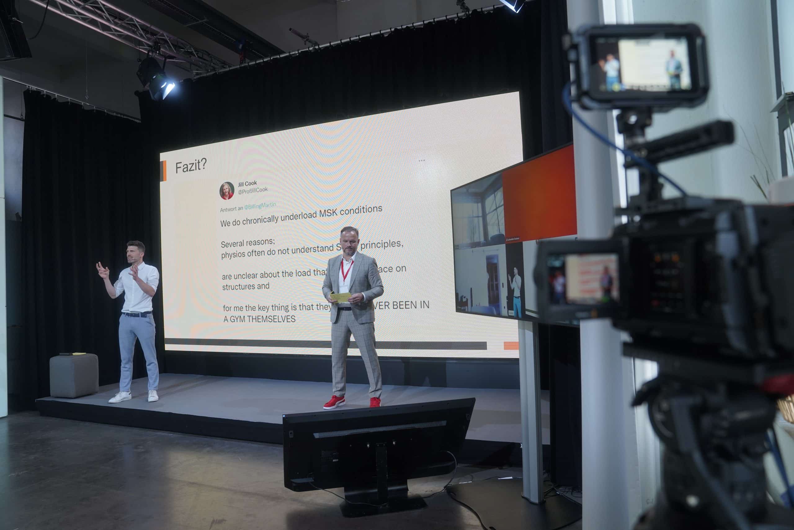 Zwei Männer auf einer Bühne bei einer Präsentation, die mit einer Kamera in das Internet übertragen wird.