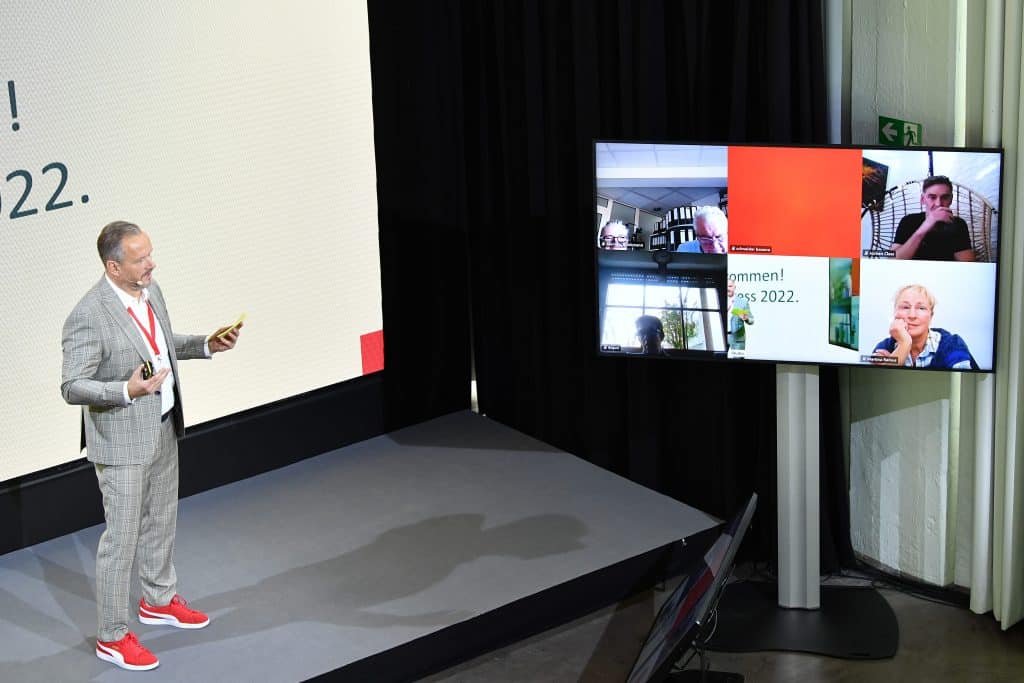 Ein man in einem grauen Anzug präsentiert auf einer Bühne bei einer hybriden Veranstaltung. Neben ihm sind auf einem Bildschirm andere Personen Online zugeschaltet.
