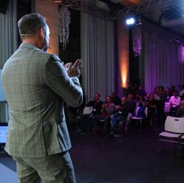 Ein Mann mit einem Headset Mikrofon auf einer Tagung präsentiert vor Publikum.
