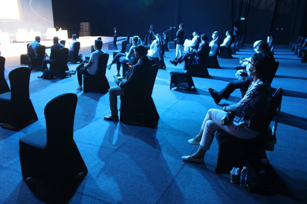 Ein großer Saal in blaues Licht getaucht. Die Besucher der Veranstaltung sitzen weit auseinander und tragen Masken.