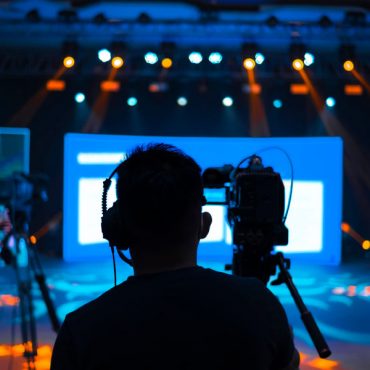 Kameramänner vor einer orange und blau ausgeleuchteten Bühne, die mit Licht und Projektionstechnik ausgestattet ist, bei der Aufzeichnung eines hybriden Event.
