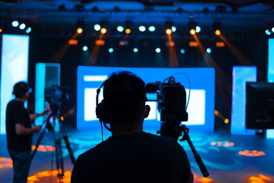Kameramänner vor einer orange und blau ausgeleuchteten Bühne, die mit Licht und Projektionstechnik ausgestattet ist, bei der Aufzeichnung eines hybriden Event.